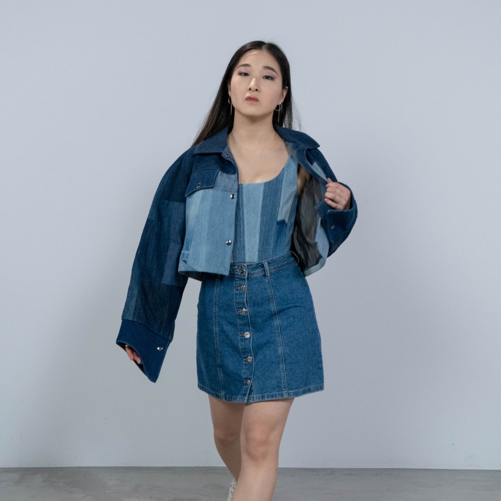A young woman in a stylish denim outfit — a denim skirt with a button-down front, a striped denim top, and a matching denim jacket — standing against a plain, light-colored background.